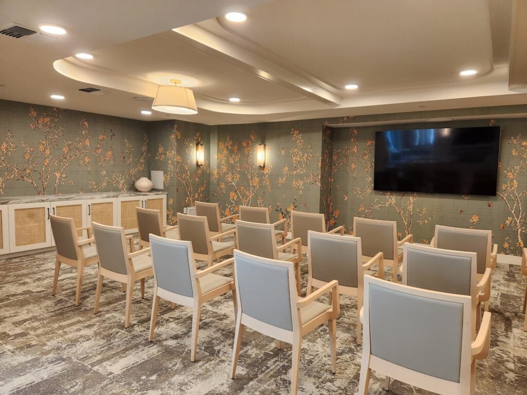 Upscale senior living room with a row of chairs and television with calming colors at The Seville